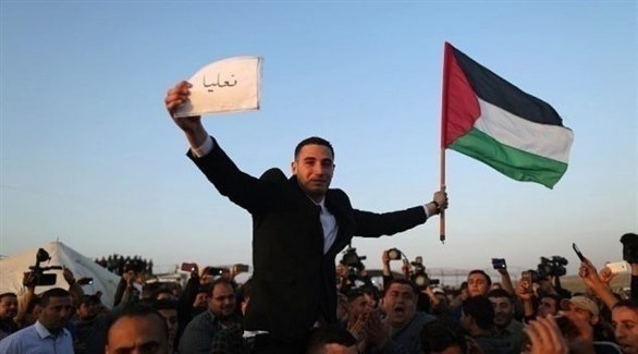 شاب يلوح بعلم فلسطيني ويحمل لافتة باسم قرية عائلته في إسرائيل على حدود غزة مع إسرائيل (أف ب)