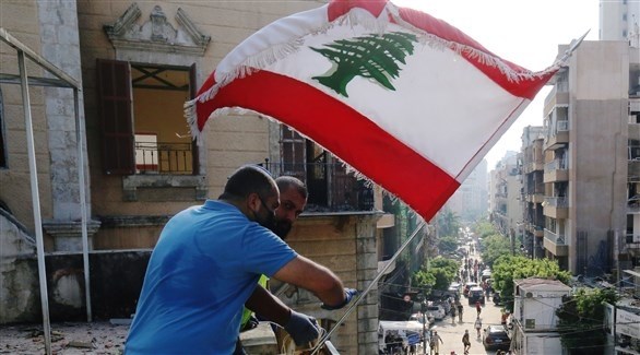 رجلان يعلقان العلم اللبناني على شرفة منزل متضرر جراء تفجير بيروت (رويترز)