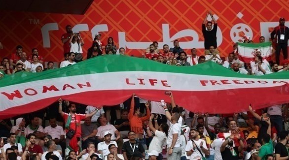 مشجعون إيرانيون يرفعون علماً بشعار الاحتجاجات في قطر (تويتر)